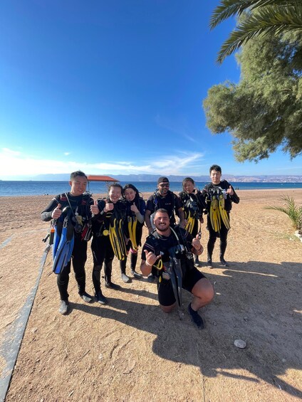 A group of people in scuba diving gear on a beach with a clear sky and mountains in the background. 2
