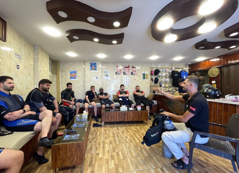 A group of people in a room with wooden flooring, some seated on benches and others standing. 4