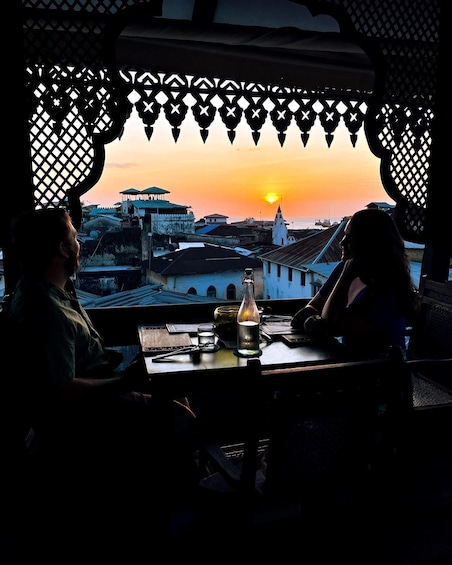 Two people seated at a table with a bottle and papers, overlooking a sunset over a coastal town. 1