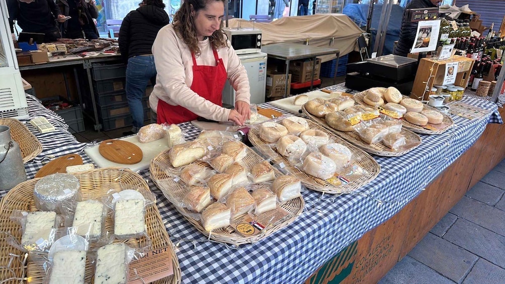 A market stall with various cheeses and breads displayed on a checkered tablecloth. 2