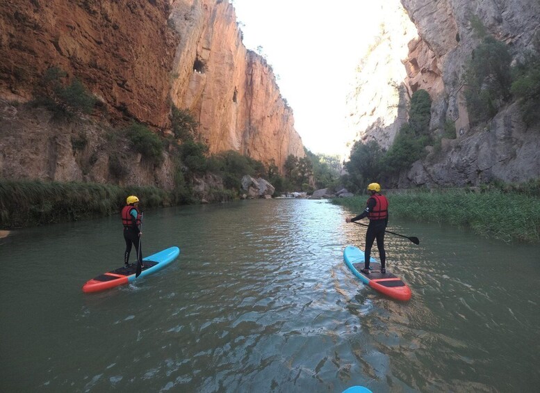 Two people paddleboarding on a river surrounded by rocky cliffs. 5