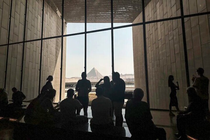 A group of people inside a building with large windows, looking out at a pyramid. 3