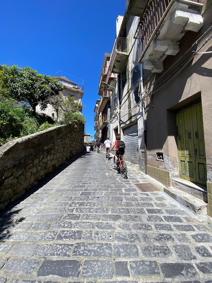 A narrow cobblestone street flanked by buildings with balconies, a stone wall on one side, and a green door on the other. 5