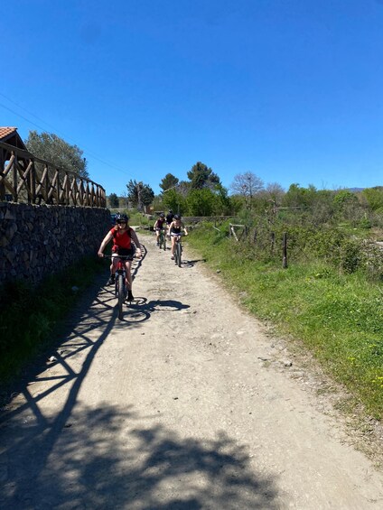 A group of cyclists on a dirt path with a stone wall and wooden fence on one side, surrounded by greenery and trees. 2
