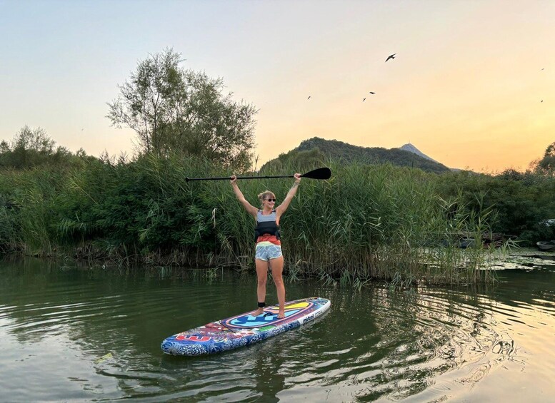 A person standing on a colorful paddleboard in a body of water, surrounded by greenery and a hill in the background. 5