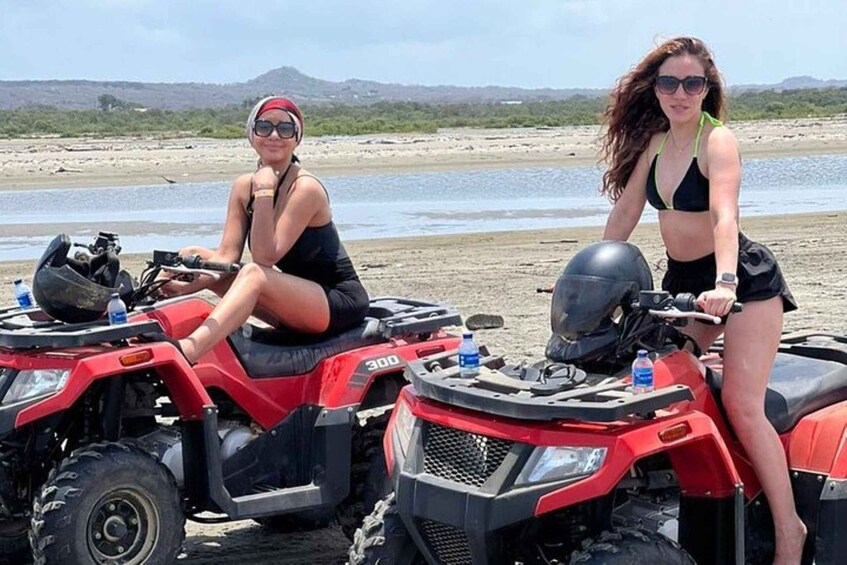 Two people on ATVs on a sandy beach with water and hills in the background. 2