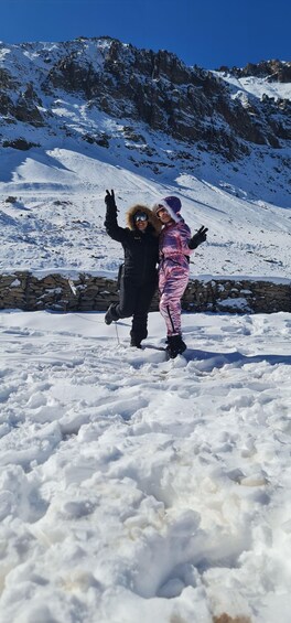 Two people in winter clothing posing on a snowy mountain with rocky terrain in the background. 3