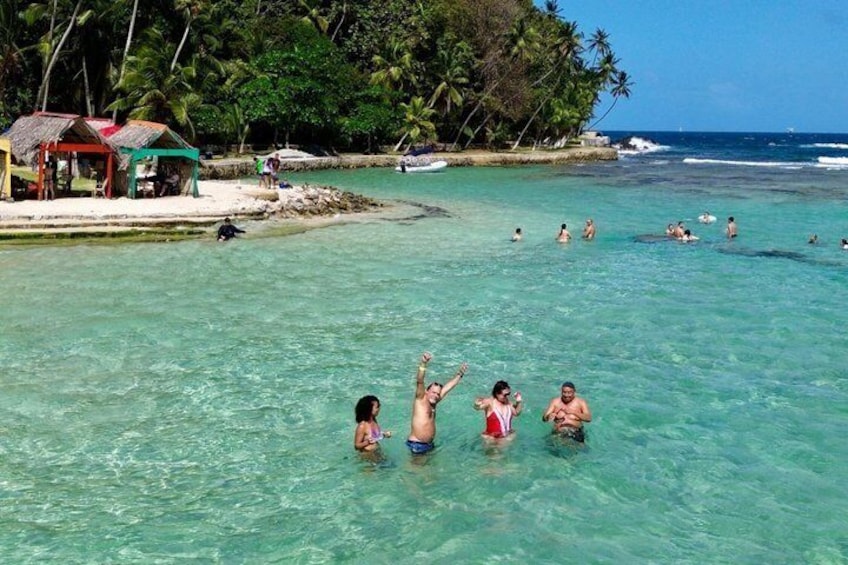 People swimming in clear turquoise water, a beach with palm trees, and a small hut. 3