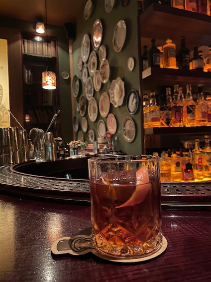 A bar with a glass of amber-colored drink, a wall of vintage plates, and shelves of liquor bottles. 2