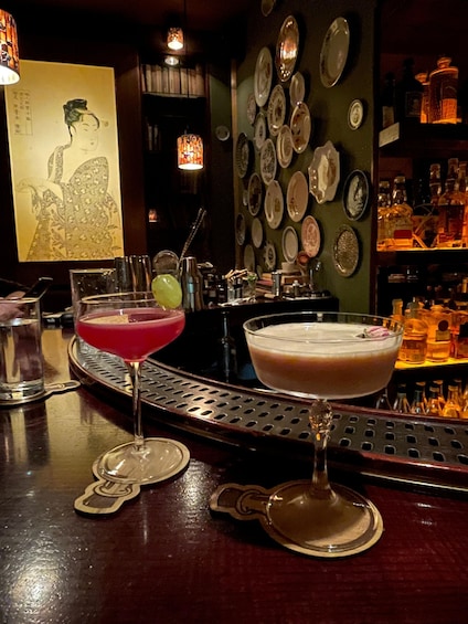 A bar with two cocktails on the counter, a decorative plate wall, and a traditional painting in the background. 1