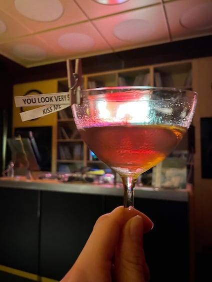 A hand holding a cocktail glass with a frothy drink, a sign above the glass, and a bar counter in the background. 4