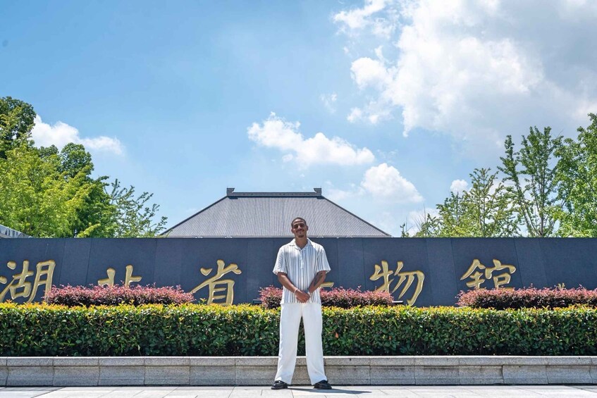 A person standing in front of a sign with Chinese characters, a building with a grey roof, and green trees in the background. 1