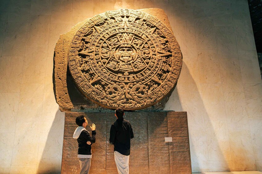 A large stone carving with intricate designs, two people observing it, and a tiled floor. 1