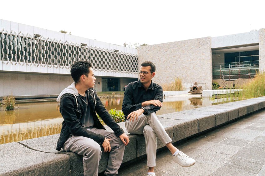 Two individuals seated on a stone bench outdoors, with a modern building and a water feature in the background. 4