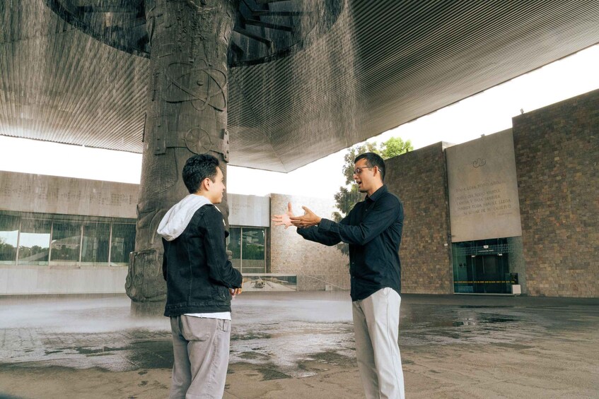 Two people in an urban setting with a large stone structure and a building with a glass facade in the background. 3