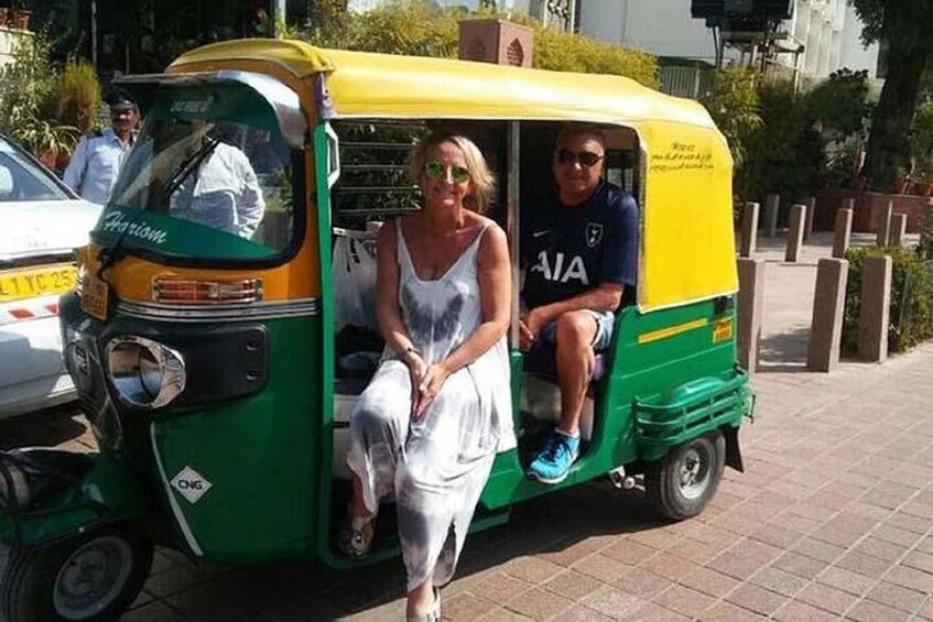 A green and yellow auto-rickshaw with two passengers, one in the driver's seat and one in the back. 1