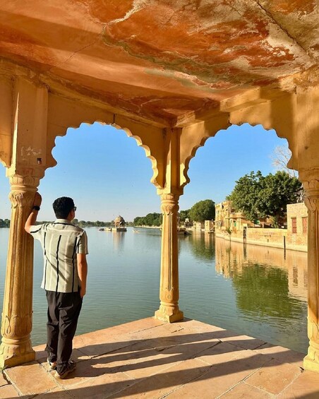 A man standing under a traditional archway by a calm river, with buildings and trees in the background. 3