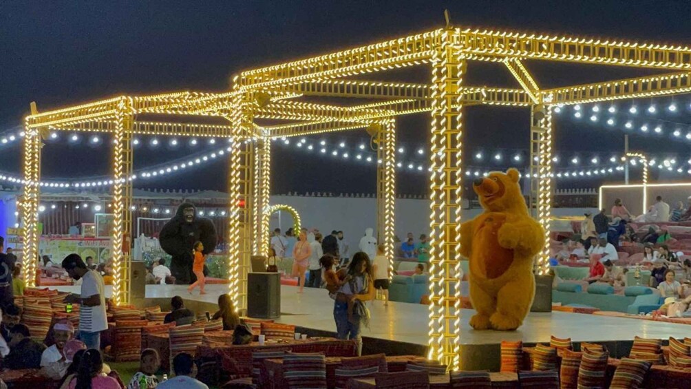 A brightly lit outdoor event with a large bear mascot, people sitting on couches, and a stage with a gorilla figure. 5
