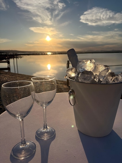 A sunset over a calm lake with a table featuring two wine glasses and a bucket of ice. 1