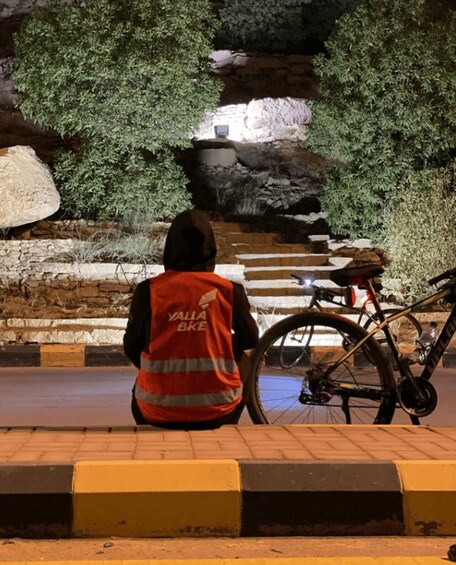 A person in a reflective vest sitting next to a bicycle on a paved area with steps leading to a stone wall and greenery. 3