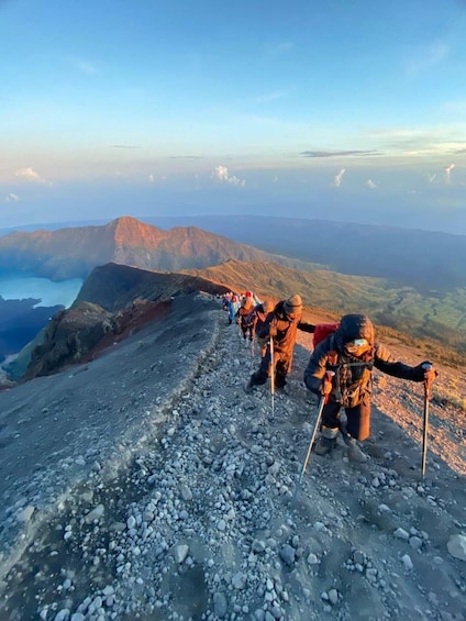 A group of hikers ascending a rocky mountain path with a panoramic view of distant mountains and a body of water. 1