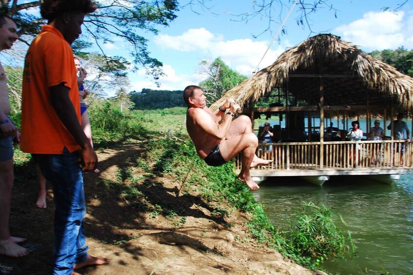 A man swinging on a rope over a river, a thatched-roof structure, and people on a dock. 2