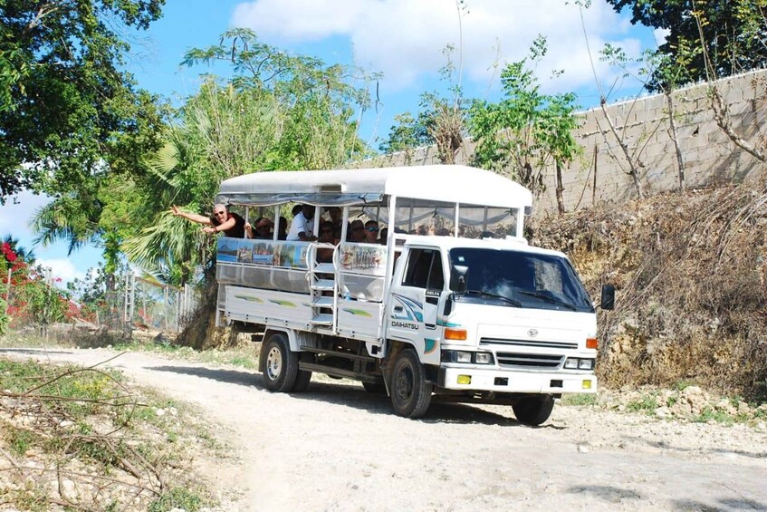 A white truck with a covered cargo area, parked on a dirt road with vegetation and a stone wall in the background. 1