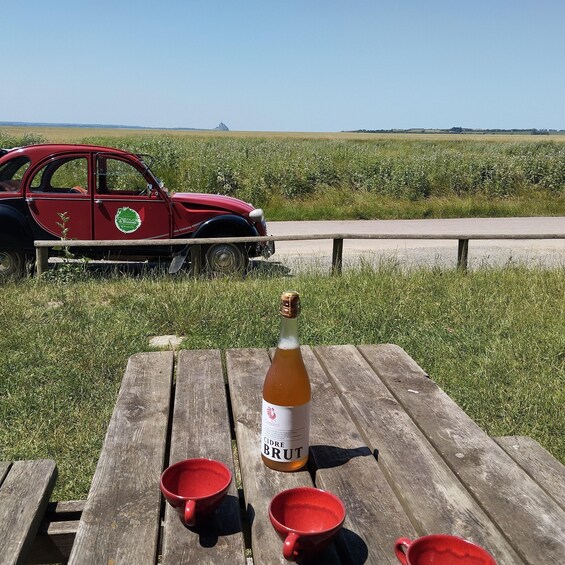 A picnic table with a bottle of cider brut and red cups, a vintage red car parked in a field. 2