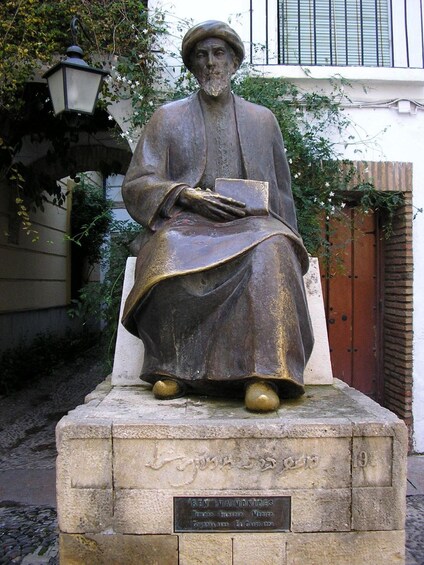 A statue of a seated man with a book, wearing traditional attire, on a pedestal with a plaque. 1