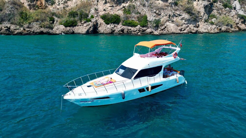 A white yacht with a canopy and a person on deck, floating on turquoise water with rocky cliffs in the background. 1