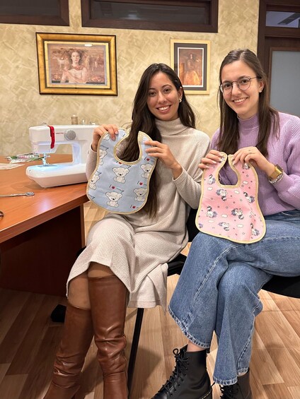 Two individuals holding baby bibs in a room with a sewing machine and framed pictures on the wall. 3