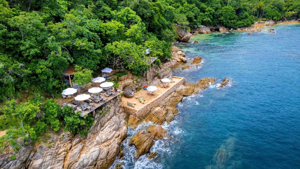 A coastal area with a rocky shoreline, lush greenery, and a series of outdoor seating areas with umbrellas. 5