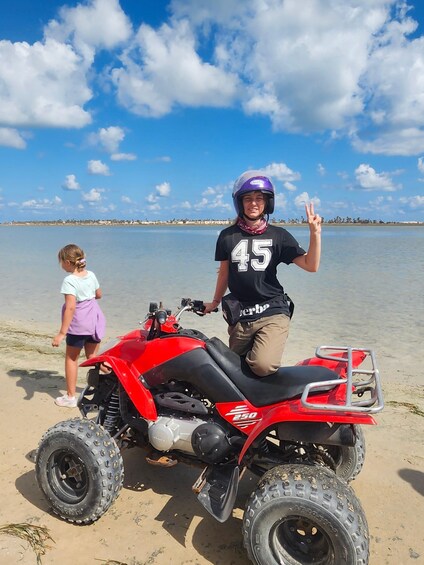 A person on a red ATV with the number 45, a child, and a beach with water in the background. 3