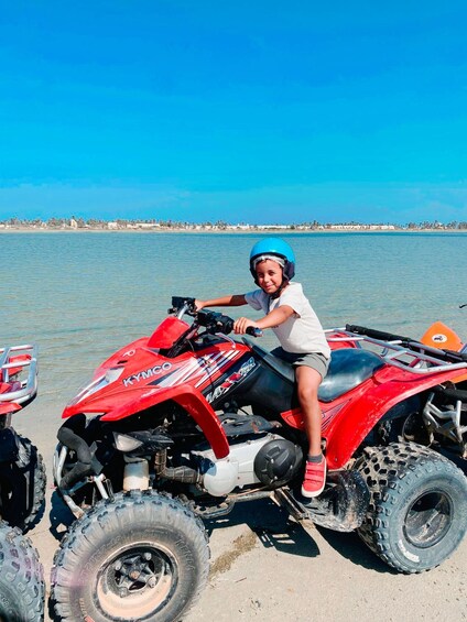 A child on a red ATV with a blue helmet, by a calm body of water with a distant shoreline. 5
