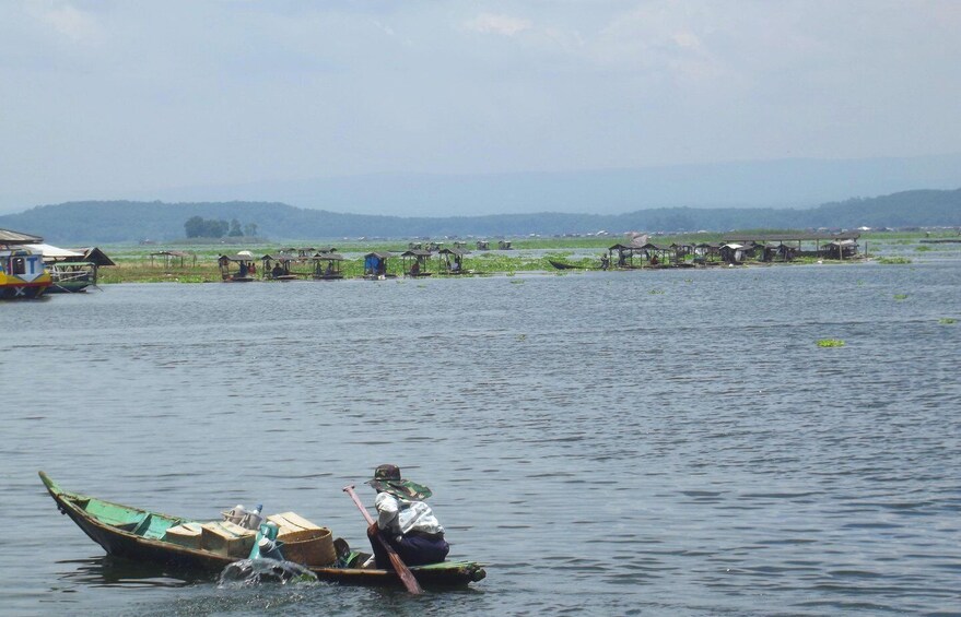 A person rowing a boat on a calm body of water with floating houses in the background. 1
