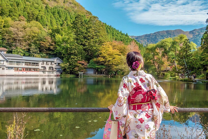 A person in a kimono standing by a calm lake with a traditional building in the background. 1