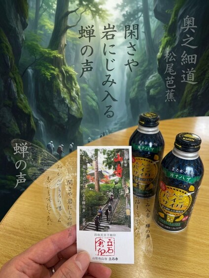 A hand holding a postcard with a forest scene, two bottles with labels, and Japanese text on a wooden table. 2