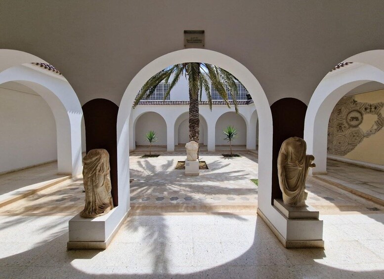 An architectural courtyard with arches, statues, palm trees, and a tiled floor. 2