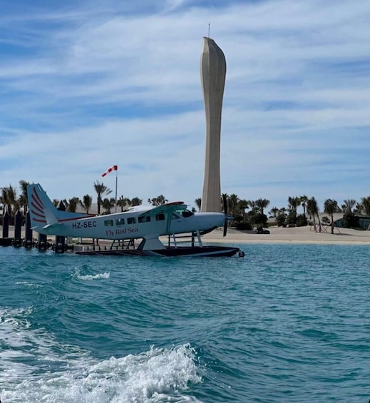 A seaplane with 'HZ-SEC' and 'Fly Red Sea' on its side, docked near a tall, slender tower by a calm sea with palm trees in the background. 1