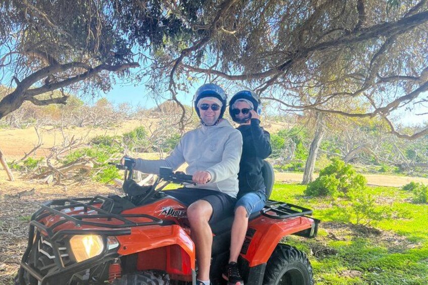 Zwei Personen fahren mit einem roten Quad durch eine natürliche Landschaft mit Bäumen und viel Grün. 2