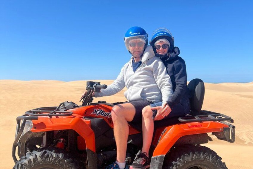 Zwei Personen fahren mit einem orangefarbenen Geländefahrzeug durch eine Wüstenlandschaft mit Sanddünen. 3