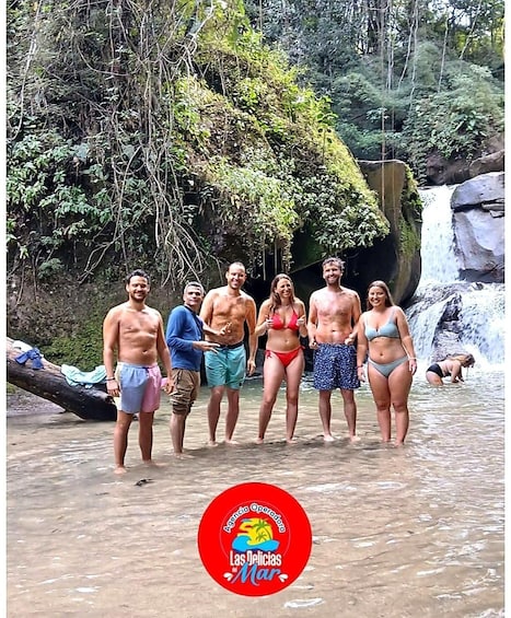 A group of people standing in shallow water near a waterfall in a forested area. 5