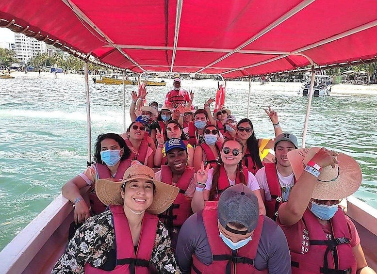 A group of people on a boat with a red canopy, wearing life jackets and some hats, on a body of water with buildings in the background. 3