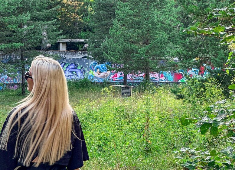 A person with long blonde hair standing in a grassy area with trees and colorful graffiti on a wall in the background. 1