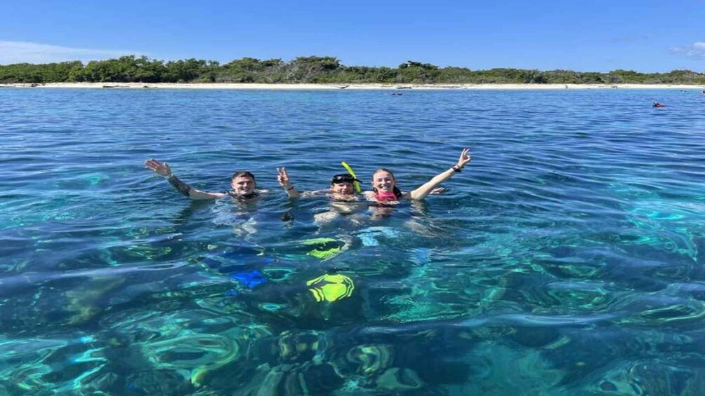 Three people snorkeling in clear blue water with a beach and vegetation in the background. 2