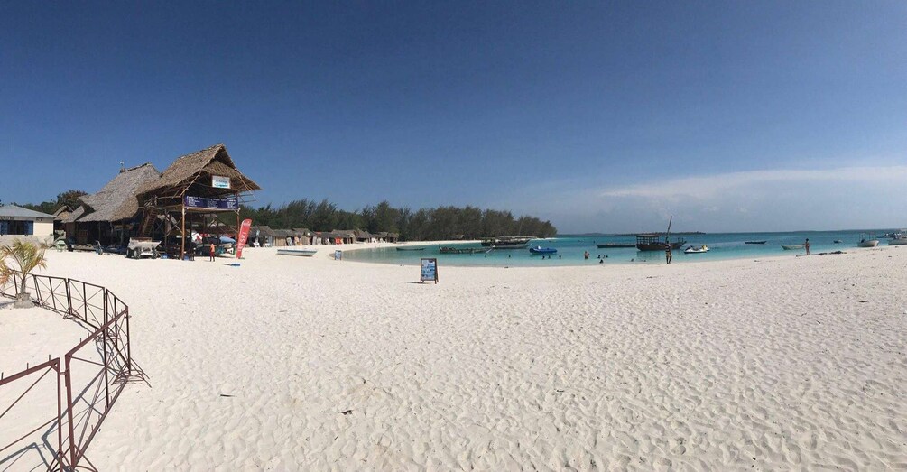 Una playa con un edificio de techo de paja, agua turquesa cristalina y varios barcos. 2