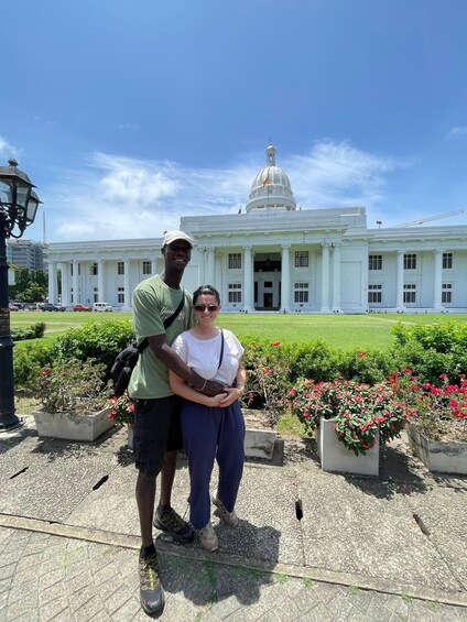 Two people standing in front of a large white building with a dome, surrounded by greenery and flowers. 4