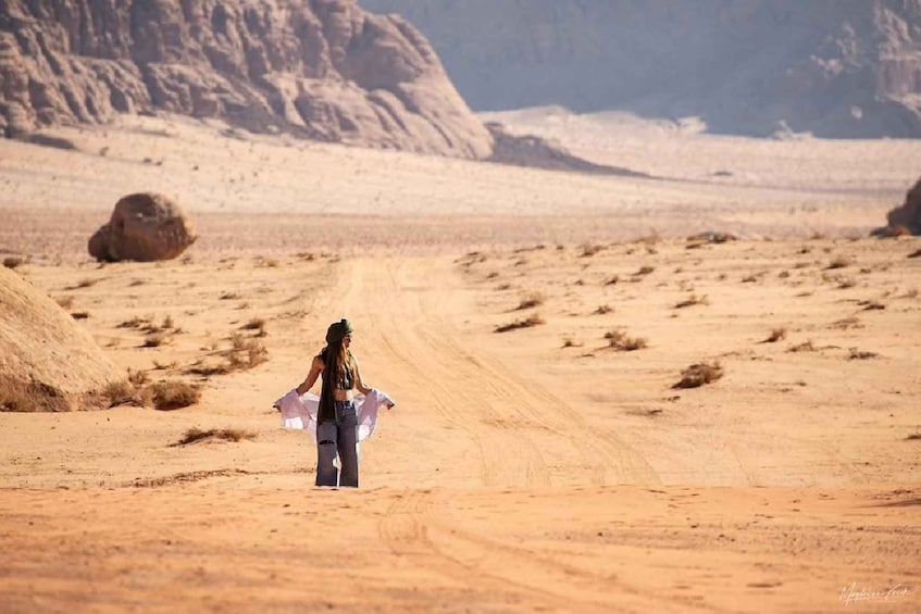 A person walking on a dirt road in a desert landscape with sand dunes and rocky formations. 3