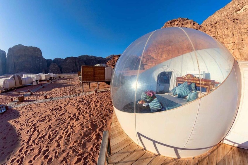 A transparent dome structure with a bed and pillows inside, set on a wooden deck in a desert landscape with rock formations. 1