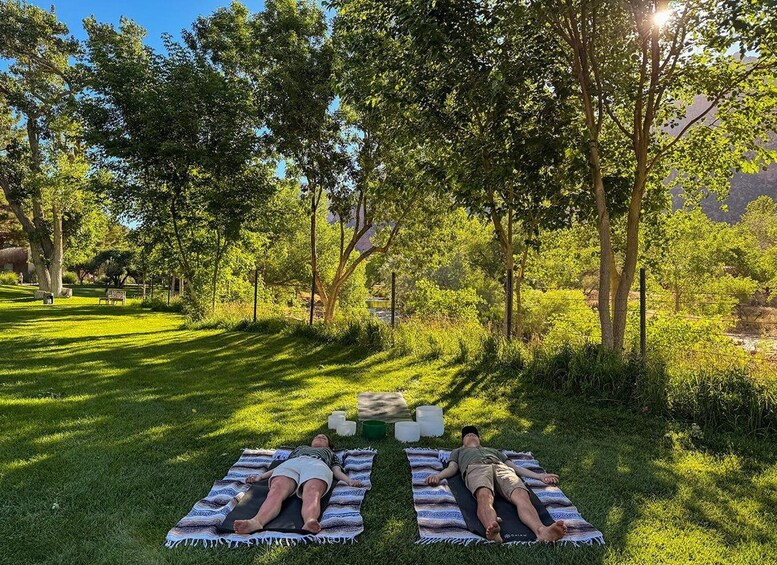 Two people lying on mats in a grassy park with trees and a bench in the background. 3
